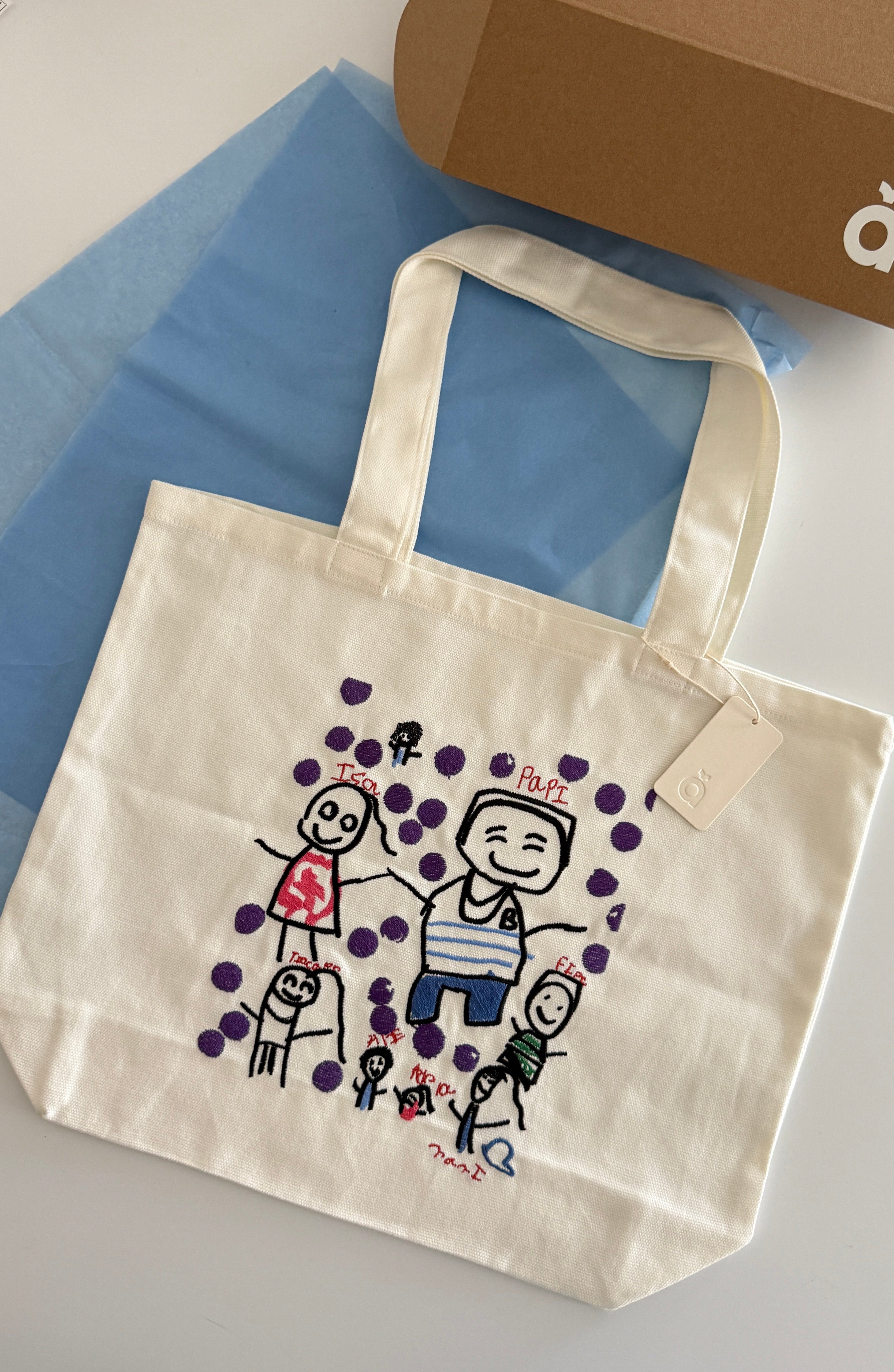 An off-white canvas tote bag with a colorful hand-drawn family illustration and purple polka dots, displayed on a light gray surface next to a brown cardboard box and blue tissue paper.