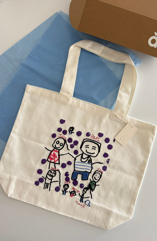 An off-white canvas tote bag with a colorful hand-drawn family illustration and purple polka dots, displayed on a light gray surface next to a brown cardboard box and blue tissue paper.